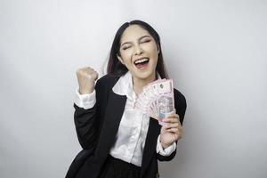 una joven mujer de negocios asiática con una expresión feliz y exitosa que usa traje negro y tiene dinero en rupias indonesias aislada de fondo blanco foto
