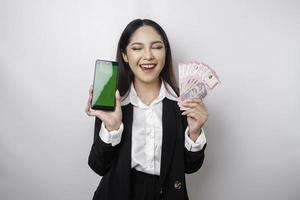 A happy young businesswoman is wearing black suit, showing her phone and money in Indonesian rupiah isolated by white background photo