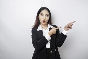 Shocked Asian businesswoman wearing a black suit pointing at the copy space beside her, isolated by a white background photo