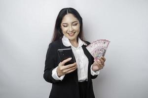 A happy young businesswoman is wearing black suit, holding her phone and money in Indonesian rupiah isolated by white background photo