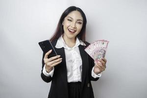 A happy young businesswoman is wearing black suit, holding her phone and money in Indonesian rupiah isolated by white background photo