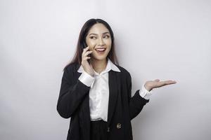 emocionada mujer de negocios asiática con traje negro apuntando al espacio de copia a su lado mientras habla por teléfono, aislada de fondo blanco foto