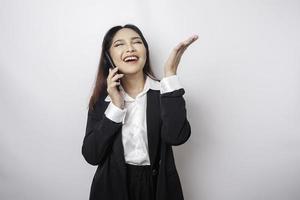 emocionada mujer de negocios asiática con traje negro apuntando al espacio de copia encima de ella mientras habla por teléfono, aislada de fondo blanco foto