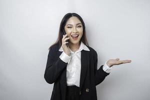 emocionada mujer de negocios asiática con traje negro apuntando al espacio de copia a su lado mientras habla por teléfono, aislada de fondo blanco foto