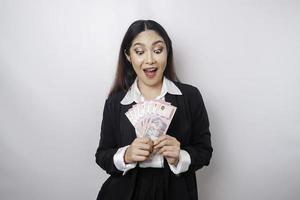 A happy young businesswoman is wearing black suit and holding cash money in Indonesian rupiah isolated by white background photo