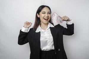 una joven mujer de negocios asiática con una expresión feliz y exitosa que usa traje negro y tiene dinero en rupias indonesias aislada de fondo blanco foto