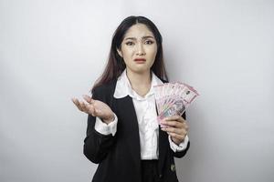 A confused young businesswoman is wearing black suit and holding cash money in Indonesian rupiah isolated by white background photo