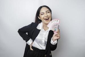 A happy young businesswoman is wearing black suit and holding cash money in Indonesian rupiah isolated by white background photo