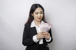 una joven empresaria confundida lleva traje negro y tiene dinero en efectivo en rupias indonesias aislada de fondo blanco foto