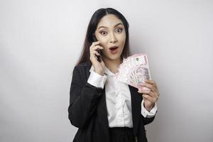 sorprendida mujer de negocios asiática con traje negro sosteniendo su teléfono inteligente y dinero en rupias indonesias, aislada de fondo blanco foto