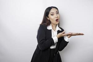 Shocked Asian businesswoman wearing a black suit pointing at the copy space beside her, isolated by a white background photo