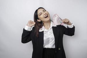 una joven mujer de negocios asiática con una expresión feliz y exitosa que usa traje negro y tiene dinero en rupias indonesias aislada de fondo blanco foto