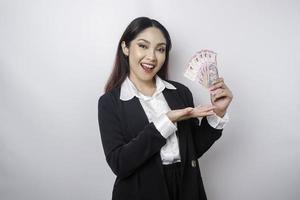 A happy young businesswoman is wearing black suit and holding cash money in Indonesian rupiah isolated by white background photo