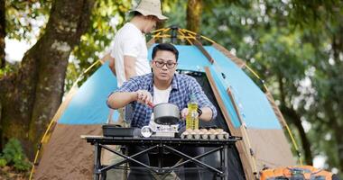 hombre asiático preparando una tienda de campaña en el campamento mientras el otro hombre preparaba comida. en acampar set de cocina al frente suelo. cocina al aire libre, viajes, camping, concepto de estilo de vida. video