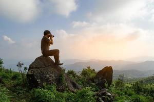 toma en gran angular de un hombre asiático sentado en la roca y toma una foto, relájate en la naturaleza y explora la luz del amanecer de la computadora conceptual, copia el espacio para el texto individual foto