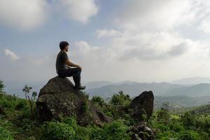 Wide angle shot of Asian man sitting on the rock and look at far a way adventure alone, nature travel and environment concept computer sunrise light, copy space for individual text photo