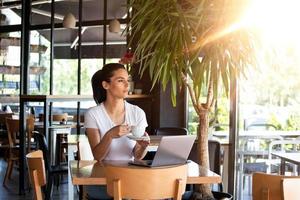 Young beautiful girl sitting at a table in a cafe by the window, drinking cappuccino coffee. Beautiful woman working with laptop from coffee shop. Attractive woman sitting in a cafe with a laptop photo