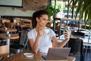 Portrait of a playful young girl taking selfie with mobile phone while sitting with laptop computer at a cafe outdoors. Morning coffee is my daily routine photo