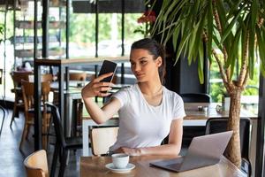 Portrait of a playful young girl taking selfie with mobile phone while sitting with laptop computer at a cafe outdoors. Morning coffee is my daily routine photo