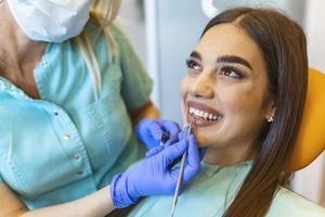 Image of satisfied young woman sitting in dental chair at medical center while professional doctor fixing her teeth, Female dentist choosing filling shade for smiling woman, using tooth scale sample photo