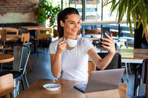 Portrait of a playful young girl taking selfie with mobile phone while sitting with laptop computer at a cafe outdoors. Morning coffee is my daily routine photo