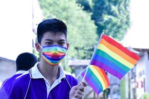 retrato de un joven asiático que usa máscara de arco iris y sostiene una bandera de arco iris, símbolo lgbt, en el parque, enfoque suave y selectivo, concepto para la celebración de la comunidad lgbt y respeto por la diversidad de género. foto