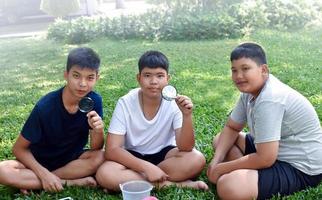 Three Asian studets holding magnifying glasses and other learning devices, tablets, small fishing nets and a small white plastic bin to learn about underwater insects after taking it out of the pond. photo