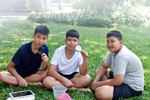 Three Asian studets holding magnifying glasses and other learning devices, tablets, small fishing nets and a small white plastic bin to learn about underwater insects after taking it out of the pond. photo