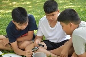Three Asian studets holding magnifying glasses and other learning devices, tablets, small fishing nets and a small white plastic bin to learn about underwater insects after taking it out of the pond. photo