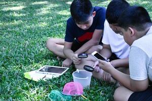 tres estudiantes asiáticos con lupas y otros dispositivos de aprendizaje, tabletas, pequeñas redes de pesca y un pequeño contenedor de plástico blanco para aprender sobre insectos submarinos después de sacarlos del estanque. foto