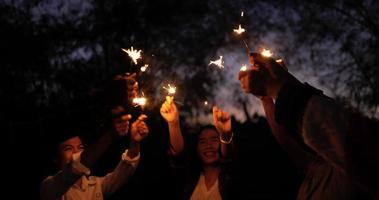 tiro panorámico de mano, grupo de jóvenes amigos sosteniendo fuego quemando bengalas, están alegres saludando para jugar con brillo en las manos, sonríen y ríen juntos en la noche de fiesta de año nuevo al aire libre video