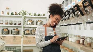 A Black female shopkeeper works at refill store, talks on mobile phone with customer checks stock orders of organic products in reusable containers, zero-waste grocery shop, and sustainable business. video