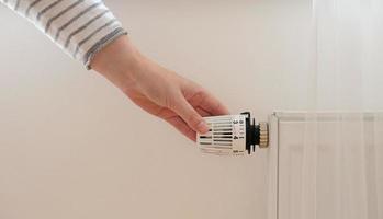 mujer regula la temperatura en casa con el termostato de calefacción para ahorrar energía, primer plano con la mano. válvula de control de temperatura en el radiador. calentador blanco en la pared con cortina en apartamento moderno. foto