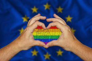 manos masculinas formando un corazón con una bandera lgbt dentro contra la bandera de fondo de europa foto