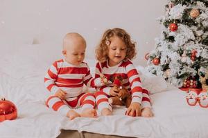 children in red and white pajamas try on funny glasses with Santa Claus sitting in bed. lifestyle. brother and sister celebrating Christmas. boy and girl are playing at home. High quality photo