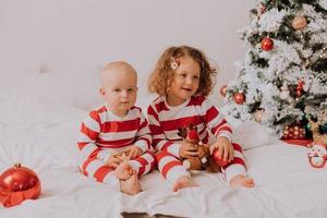 children in red and white pajamas try on funny glasses with Santa Claus sitting in bed. lifestyle. brother and sister celebrating Christmas. boy and girl are playing at home. High quality photo