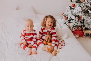 children in red and white pajamas try on funny glasses with Santa Claus sitting in bed. lifestyle. brother and sister celebrating Christmas. boy and girl are playing at home. High quality photo