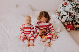 children in red and white pajamas try on funny glasses with Santa Claus sitting in bed. lifestyle. brother and sister celebrating Christmas. boy and girl are playing at home. High quality photo