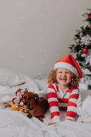 little girl in striped pajamas and a Santa hat and dog in funny glasses with Santa Claus are lying in bed on a white sheet against the background of Christmas tree. space for text. High quality photo