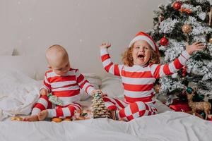 children in red and white pajamas try on funny glasses with Santa Claus sitting in bed. lifestyle. brother and sister celebrating Christmas. boy and girl are playing at home. High quality photo