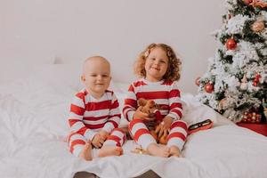 children in red and white pajamas try on funny glasses with Santa Claus sitting in bed. lifestyle. brother and sister celebrating Christmas. boy and girl are playing at home. High quality photo