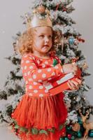 little curly-haired girl in a carnival dress hid her face behind shiny Christmas tree toys stars. child in a red dress with a Santa print on the background of a Christmas tree. High quality photo