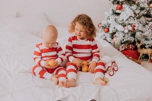 children in red and white pajamas try on funny glasses with Santa Claus sitting in bed. lifestyle. brother and sister celebrating Christmas. boy and girl are playing at home. High quality photo