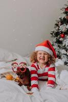 little girl in striped pajamas and a Santa hat and dog in funny glasses with Santa Claus are lying in bed on a white sheet against the background of Christmas tree. space for text. High quality photo