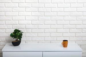 Cup and green pot on a white table near the white wall. Empty workplace in the room. White background mock up. photo