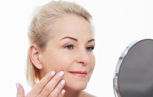 Happy middle aged woman model touching face skin looking in mirror. Smiling mature older lady enjoying healthy skin care, aging beauty, skincare treatment cosmetic products concept. photo