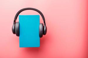 Headphones are worn on a book in a blue hardcover on a pink background, top view. photo