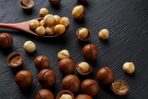 nuez de macadamia sin cáscara y nuez de macadamia pelada sobre un fondo de textura negra en una cuchara de madera foto