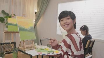 uno asiatico alunno ragazzo con pennello e tavolozza giri indietro con sorrisi, acrilico colore pittura su tela nel arte aula creativo apprendimento con talenti abilità a elementare scuola studio formazione scolastica. video