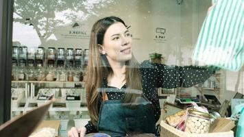 Young Caucasian female shopkeeper works by cleaning glass window displays for shop openings at refill store, zero waste, plastic-free grocery merchandise, and an eco-friendly retail business startup.
